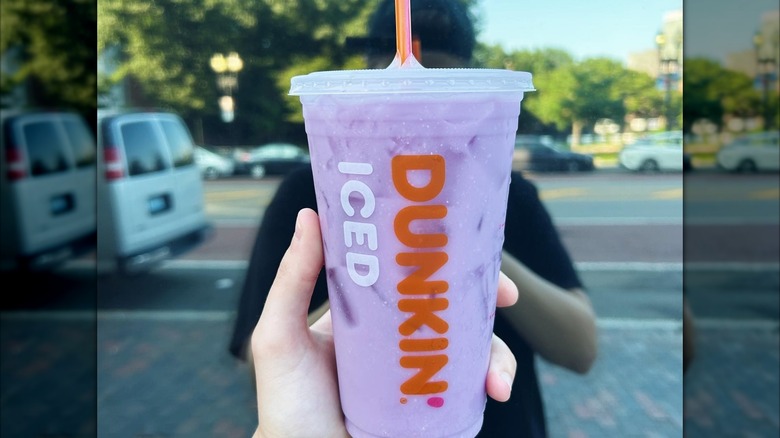 An iced raspberry watermelon Dunkin' coconut refresher held up in front of a street